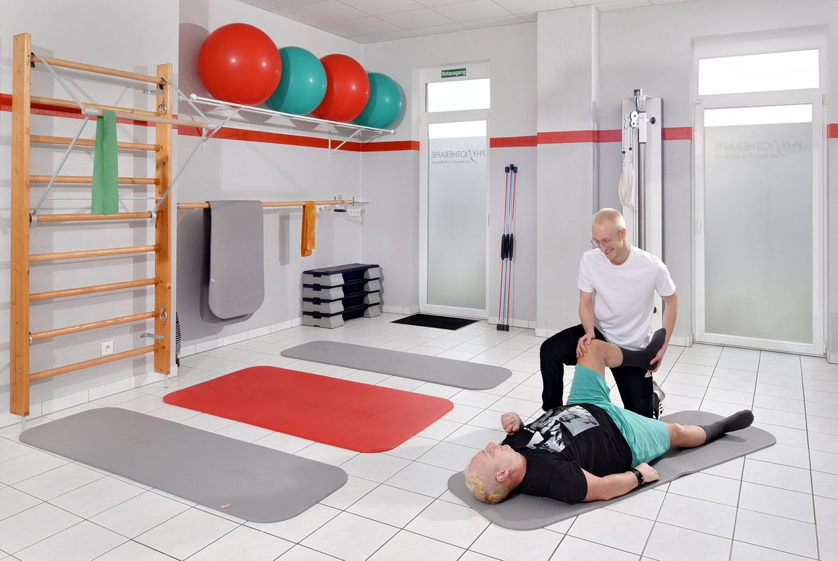 Heller Trainingsraum mit Sprossenwand, Gymnastikb&auml;llen und Matten, ein Therapeut mobilisiert das Knie eines Patienten